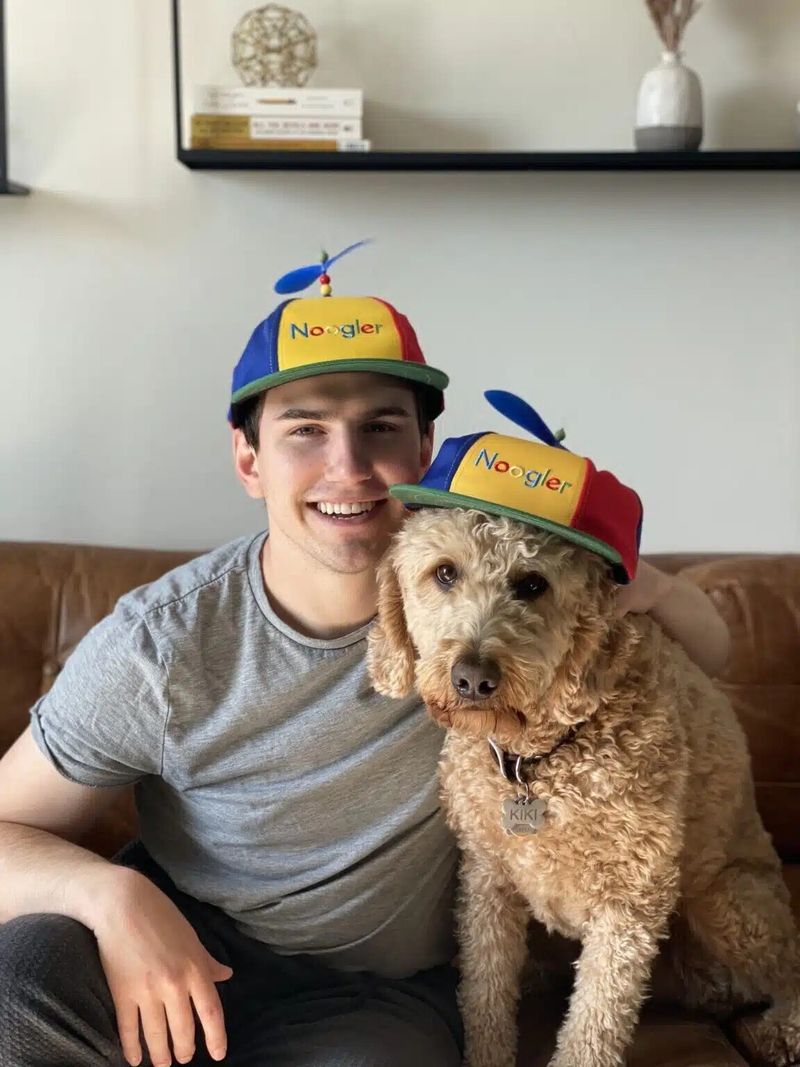 Smiling photo of a bootcamp grad wearing a Noogler (new Googler) hat. Beside him is a goldendoodle wearing a Noogler hat.